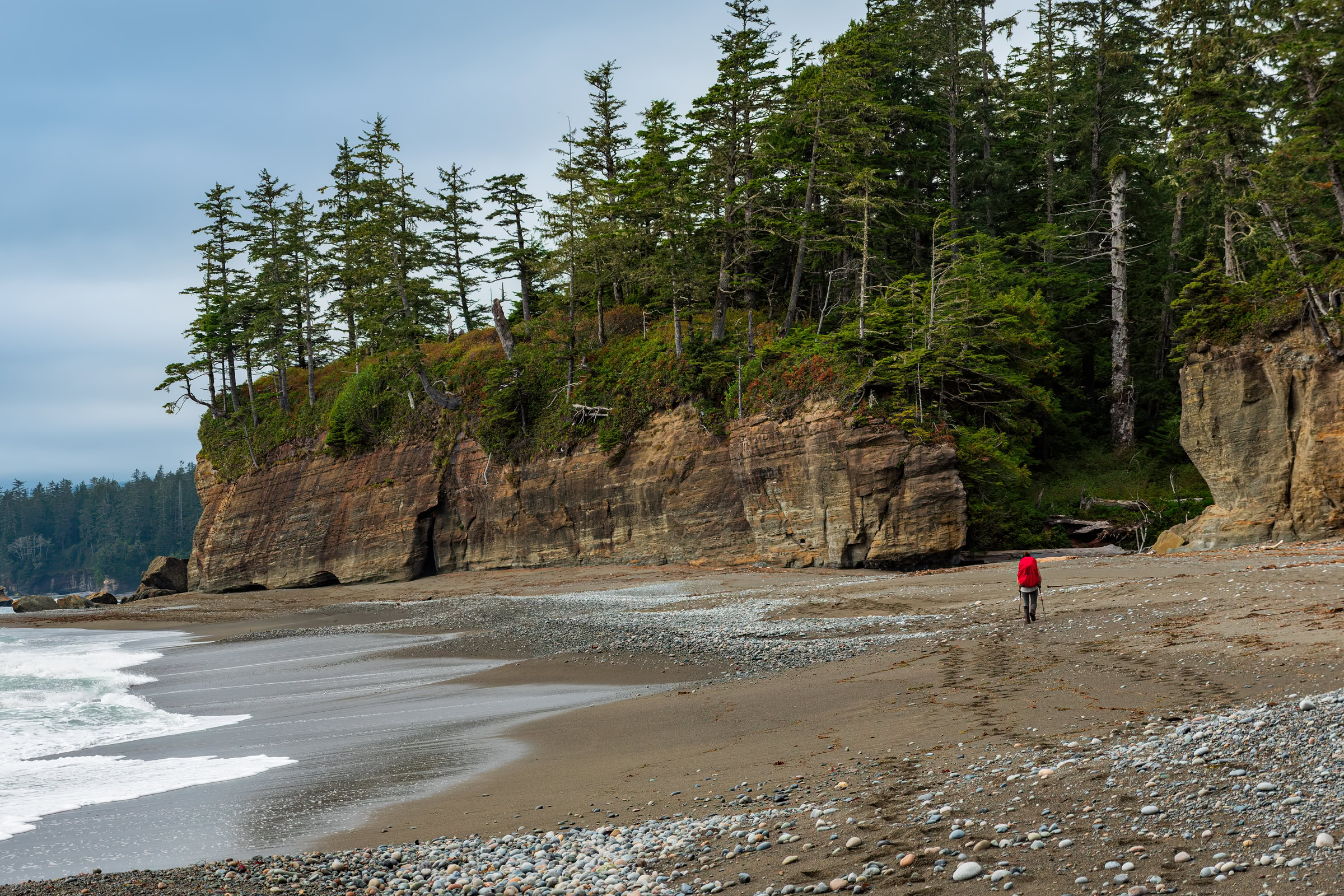 West Coast Trail wall art showing a hiker with a red pack crossing a sand and pebble beach below layered cliffs near Tsusiat Falls, Vancouver Island, BC, by James Wheeler.