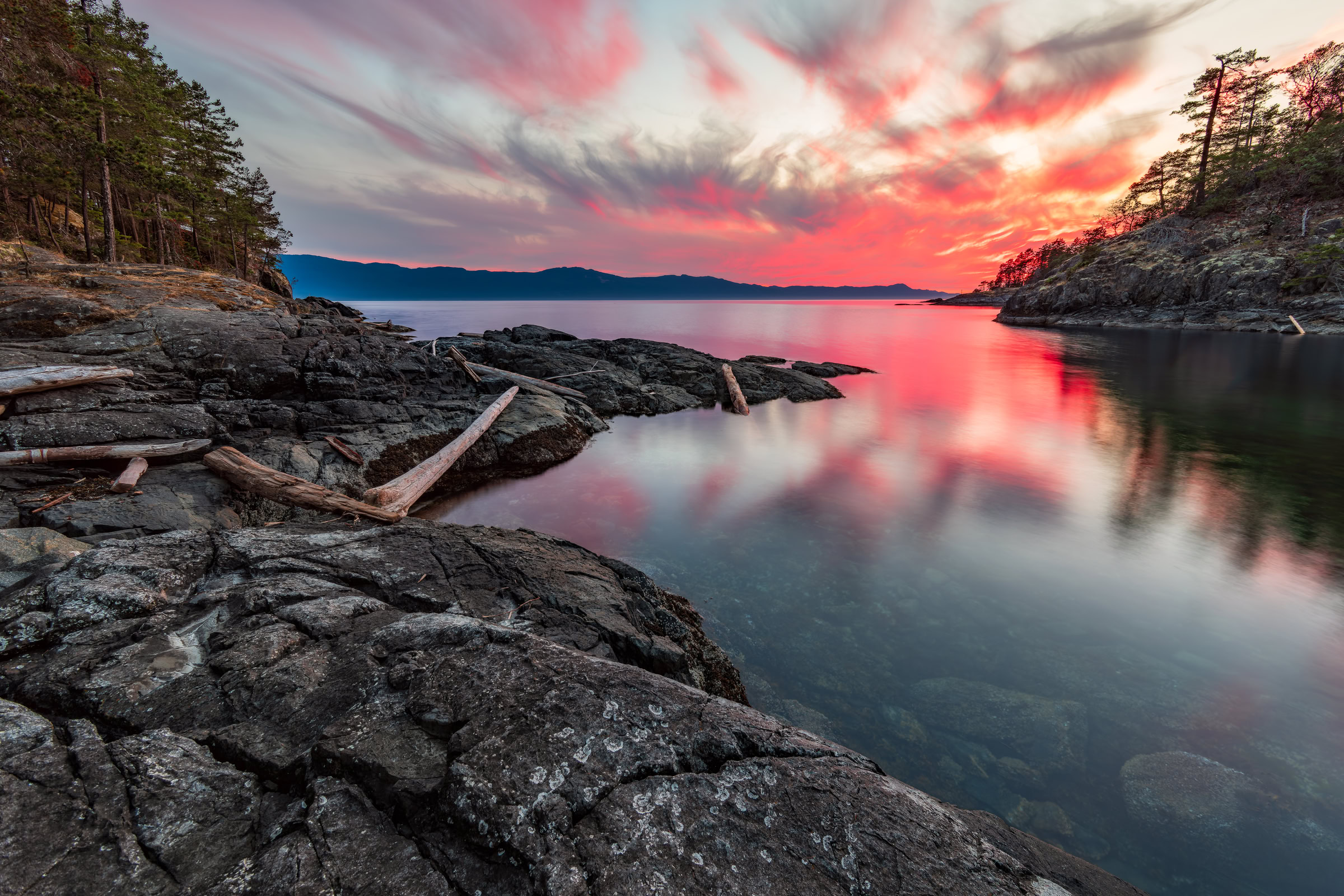 Sunshine Coast BC sunset wall art of pink clouds mirrored in calm Middle Bay beside driftwood on dark rock at Francis Point, BC, by James Wheeler.
