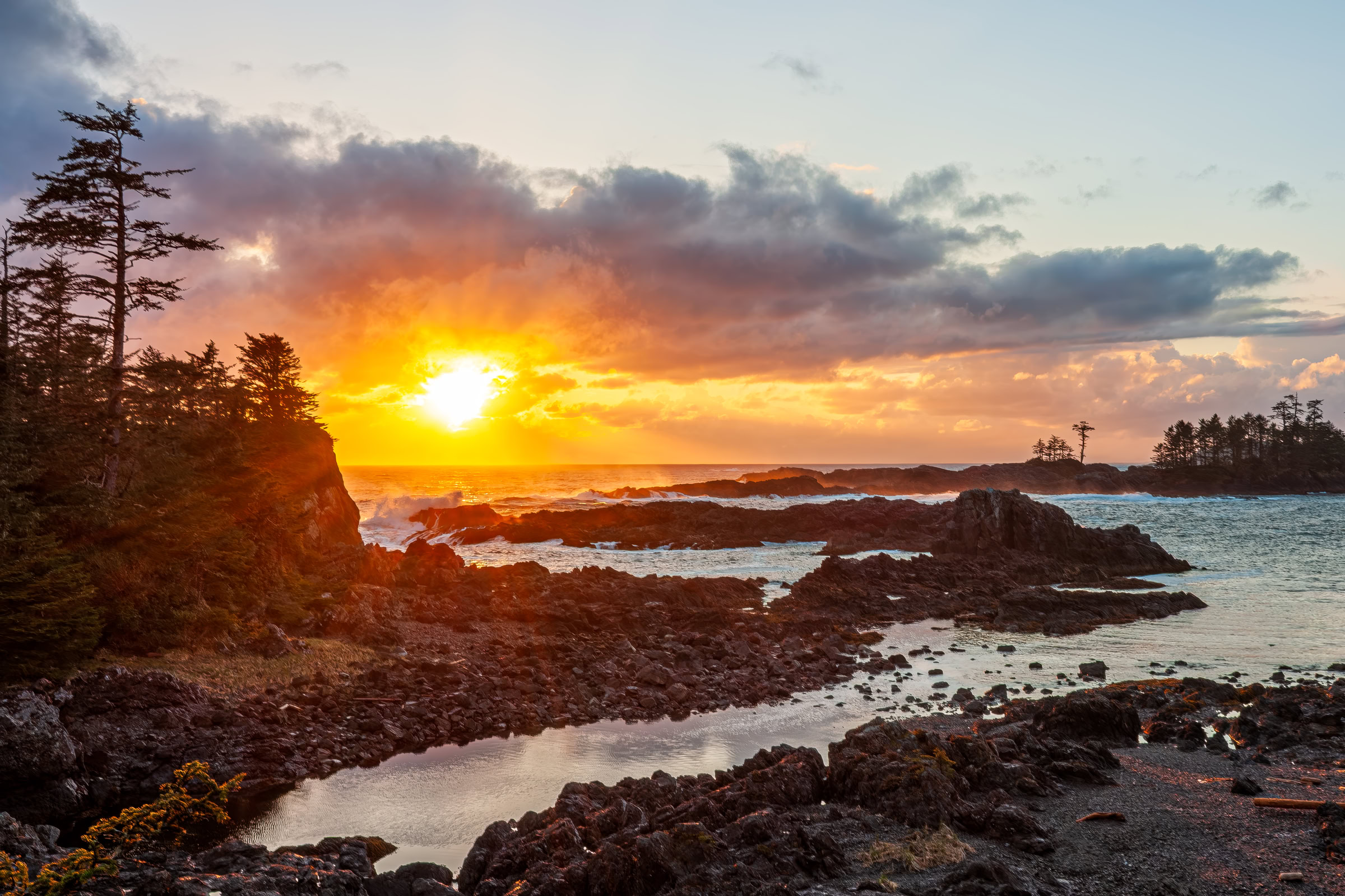 Ucluelet wall art of the sun breaking through clouds at sunset over the Pacific, waves on shore rock below the Wild Pacific Trail, BC, by James Wheeler.
